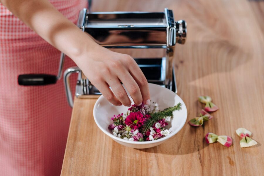 A woman decorates a pasta dish with edible flowers using a pasta maker on a wooden board.